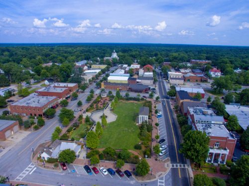 Aerial view of a small town with green spaces, buildings, and a clear blue sky in the background.