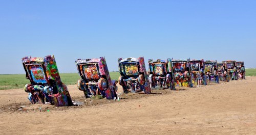 A row of colorful, graffiti-covered cars partially buried in the ground against a clear blue sky.