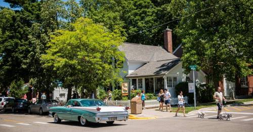 A vintage car drives past a busy street with people walking and trees lining the road on a sunny day.