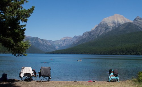 Two camping chairs by a serene lake, surrounded by mountains and clear blue skies. A small boat is visible on the water.