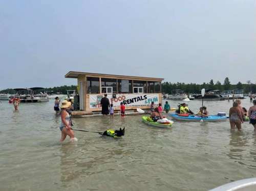 A busy beach scene with people in the water, a food rental stand, and kayaks, with a dog being walked nearby.