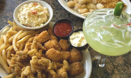 A plate of fried seafood with fries, coleslaw, dipping sauces, and a green cocktail with a lime garnish.
