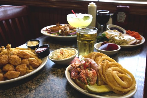A table set with various dishes: fried seafood, onion rings, coleslaw, drinks, and condiments in a casual dining setting.