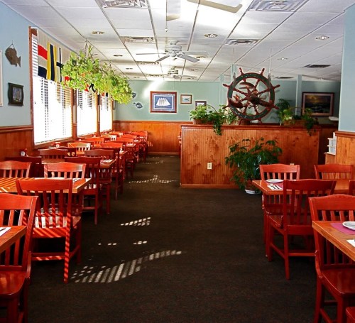 Cozy restaurant interior with wooden tables, chairs, plants, and nautical decor, featuring large windows and a ship's wheel.
