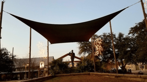 A large shade sail above a park area, with a statue and festive lights in the background during twilight.