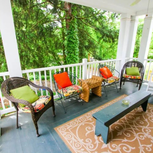 A cozy porch with wicker chairs, colorful cushions, and a small table, surrounded by lush greenery.