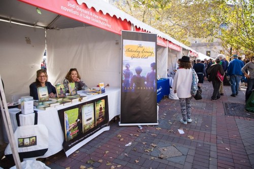 Two women sit at a book stall with various titles displayed, while people browse nearby at an outdoor event.