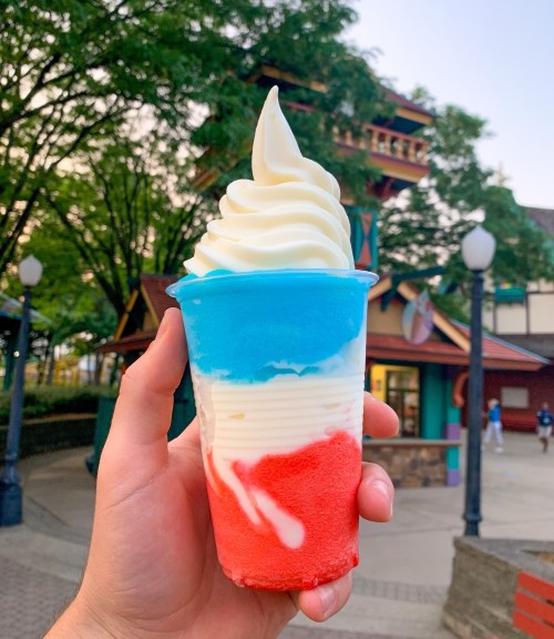 A hand holding a colorful ice cream cup with red, white, and blue layers topped with soft serve.