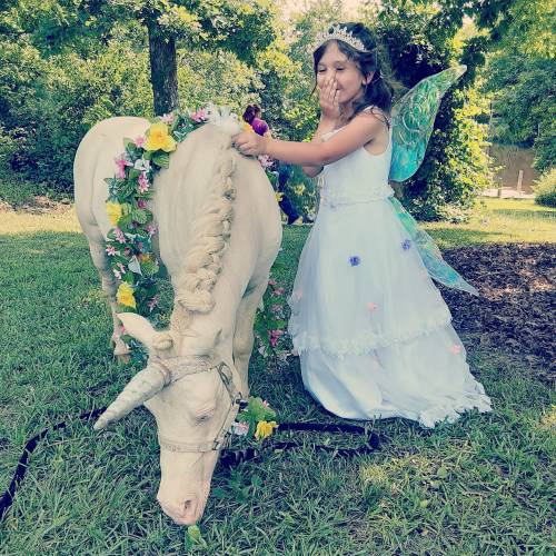 A girl in a fairy costume decorates a white unicorn with flowers in a lush, green outdoor setting.