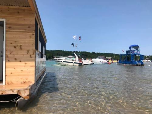A sunny day at the lake with boats anchored near a wooden houseboat and a colorful floating structure in the background.