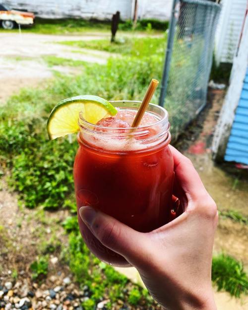 A hand holding a mason jar filled with a red drink, garnished with a lime slice and a straw, set against a grassy background.