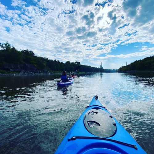 A view from a kayak on a calm river, with two kayakers in the distance under a blue sky with scattered clouds.