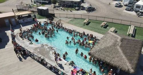 Aerial view of a crowded pool area with people swimming and lounging, surrounded by RVs and grassy spaces.