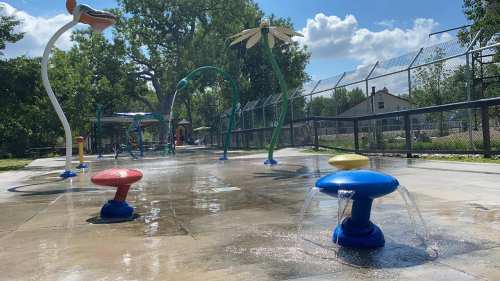 Colorful water features and spray elements in a sunny splash pad area, surrounded by greenery and a fence.