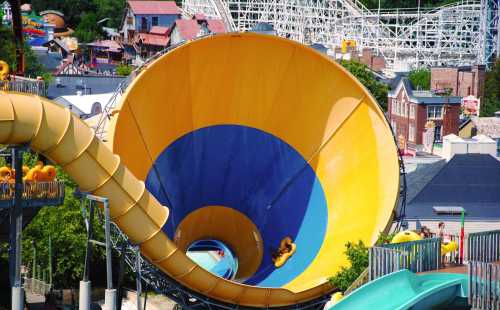 A colorful water slide spirals into a large funnel, with a rider inside, surrounded by amusement park attractions.