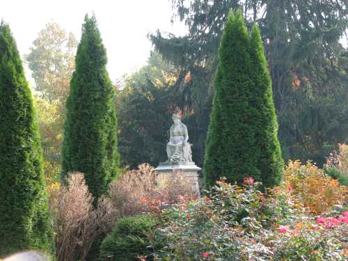 A statue seated among tall evergreen trees and colorful foliage in a serene garden setting.