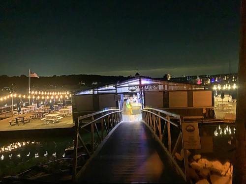 A wooden walkway leads to a lit building by the water at night, with string lights and picnic tables visible.