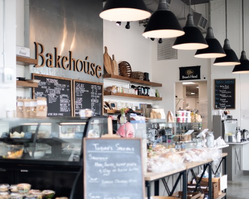 A cozy bakery interior with a display case, wooden shelves, and a sign reading "Bakehouse" under pendant lights.