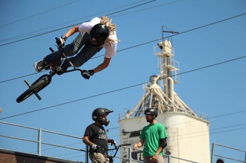 A BMX rider performs a jump at a skate park, with onlookers and a industrial building in the background.