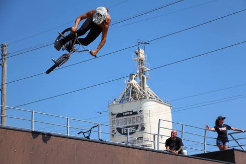A BMX rider performs a jump over a ramp, with spectators watching and a grain silo in the background under a clear blue sky.