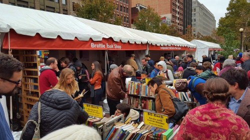 A bustling outdoor book fair with people browsing books at tents, surrounded by trees and city buildings.