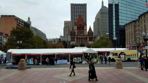 A bustling city square with tents for an event, surrounded by tall buildings and a historic church in the background.