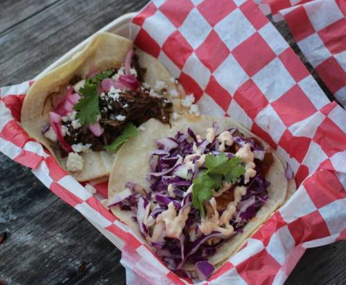 Two tacos in a red and white checkered basket: one with shredded meat, onions, and cheese; the other with purple cabbage and sauce.