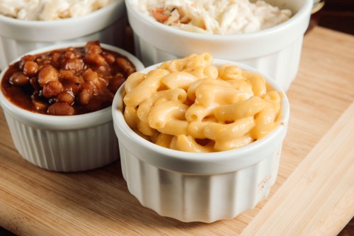A close-up of creamy macaroni and cheese in a white ramekin, alongside bowls of coleslaw and baked beans on a wooden surface.
