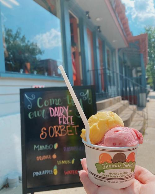 A hand holds a cup of colorful sorbet in front of a sign advertising dairy-free options at Thomas's Sweet.