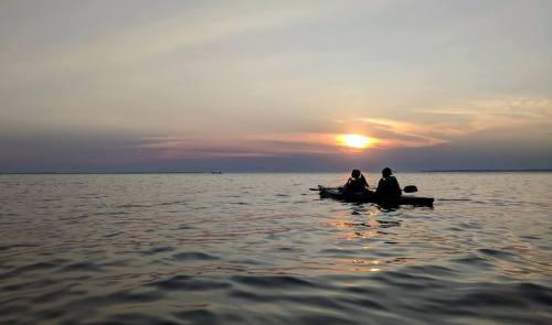 Two people kayaking on a calm lake at sunset, with soft colors reflecting on the water.