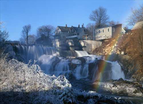 A winter scene of a waterfall with frosted trees and a rainbow, alongside a quaint house in the background.