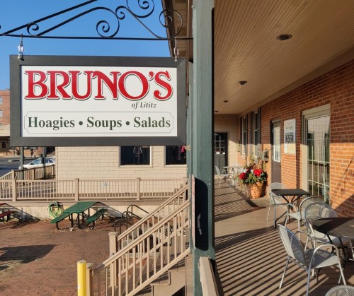 Sign for Bruno's of Lititz, featuring "Hoagies, Soups, Salads" outside a brick building with outdoor seating.
