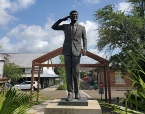 A bronze statue of a man in a suit saluting, set in a town square with buildings and trees in the background.