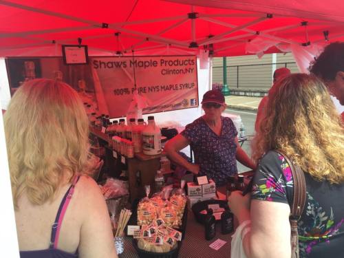 A vendor at a market booth selling Shaw's Maple Products, featuring bottles of maple syrup and snacks under a red tent.