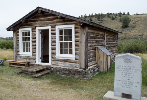 Log cabin with white windows, a small porch, and a stone monument nearby, marking the site of Esther Morris's office.