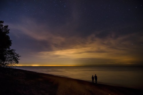 Two silhouetted figures stand on a beach under a starry night sky, with calm water and distant lights on the horizon.