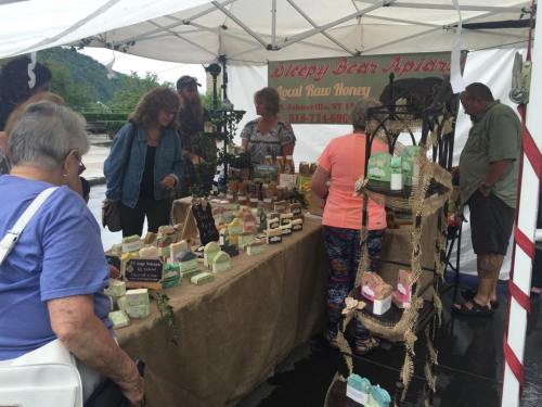 A busy market scene with people browsing a stall selling honey and handmade soaps under a white tent.