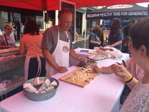 A vendor prepares food at a market under a red tent, with customers browsing in the background.