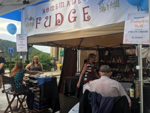 A vendor booth at a fair selling homemade fudge and pumpkin rolls, with customers browsing and enjoying treats.