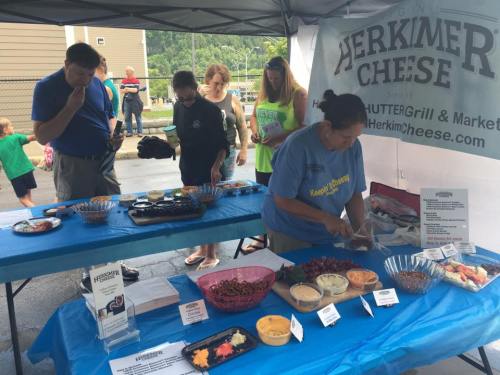 A group of people sampling food at a market booth featuring Herkimer Cheese, with various dishes displayed on a table.