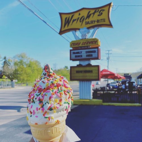A hand holds a colorful ice cream cone topped with sprinkles in front of Wright's Dairy-Rite sign on a sunny day.