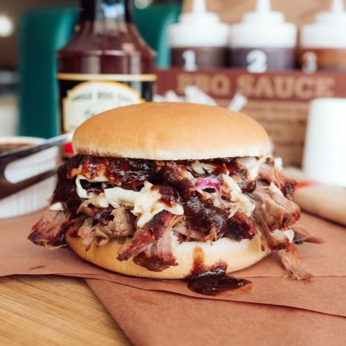A close-up of a pulled pork sandwich with coleslaw and barbecue sauce, served on a bun with condiments in the background.