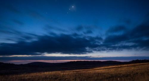 A serene twilight landscape with a darkening sky, scattered stars, and wispy clouds over a grassy field.