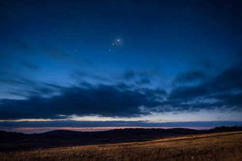 A serene night sky with stars and planets visible above a dark, grassy landscape under a twilight horizon.