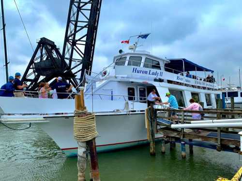 A white boat named "Huron Lady II" docked at a marina, with people boarding and a crane in the background.