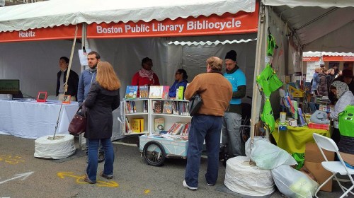 A tent with the Boston Public Library Bibliocycle, featuring books and people browsing in a lively outdoor setting.
