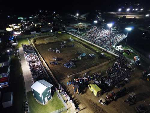 Aerial view of a nighttime event with a large crowd watching vehicles in a dirt arena, surrounded by lights and stalls.