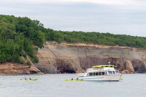 A boat and two kayakers on calm water near a rocky shoreline with lush green trees in the background.
