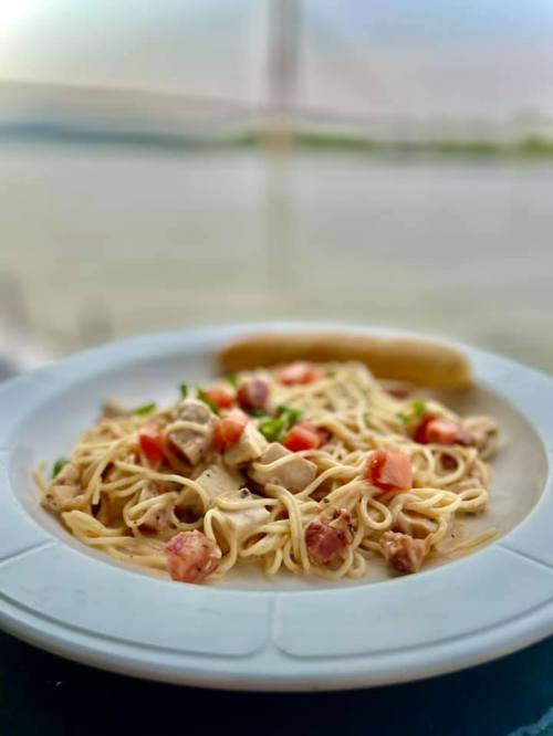 A plate of pasta with chicken, tomatoes, and herbs, set against a scenic waterfront background.