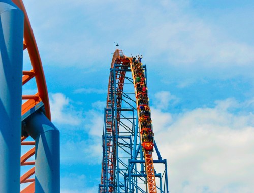 A roller coaster climbs steeply against a blue sky, with riders raising their hands in excitement.